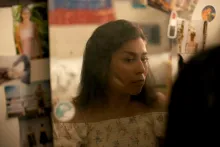 A young woman looks at herself in a bedroom mirror framed by photographs and stickers. On her cheek are two strokes of skin-toned foundation, one light and one darker than her skin tone.