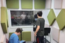 In a small room lined with green and beige-colored square panels on the walls, a person stands in front of a microphone, facing others through a control window.