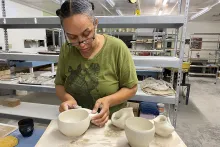 A Black woman forms a clay bowl with her hands. Other dishes in progress sit on the table in the foreground.