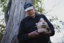 An older Black man holds up a ceramic jug shaped like a human face.