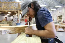 A young man fine tunes a cut in a piece of lumber using a hand tool that looks like a wide chisel.