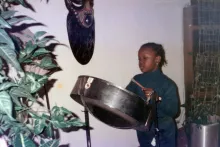 A young girl plays a steelpan drum, houseplants in the foreground and background, and a wooden mask on the wall.