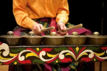 A person (face unseen) holding two wooden beaters plays a musical instrument of several gongs facing upward, sitting in a wooden frame that is painted in black, green, yellow, red, and white.