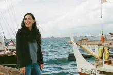 A woman with long, dark hair looks over her shoulder, out of frame. Just behind her is a ship with its name printed in black letters on the white hull: Hokule‘a.