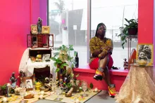 A Black woman with yellow and black printed blouse and fat sneakers sits in a window sill among pieces of an art installation, an altar with framed photos, a Black Madonna figurine, green potted plants, crystal glasses, and more.