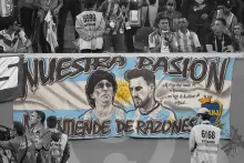 Banner in a soccer stadium with portraits of Maradona and Messi over the colors of the Argentina flag. The banner is in color, while the seated fans behind and film crew in front are in black and white.