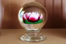 A clear glass paperweight in the shape of an orb on a pedestal, with a 3D pink-petaled rose with green leaves designed in the middle.