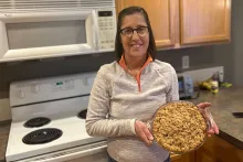 A woman with short, straight brown hair and glasses holds up a pie in a glass dish, standing in a kitchen.