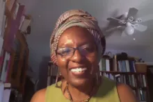 Diana N'Diaye smiles at the camera in a screenshot of a virtual conversation. She wears a earthen green tank top, glasses, an African head wrap, geometric earrings, and a wooden beaded necklace. Behind her are shelves full of books.