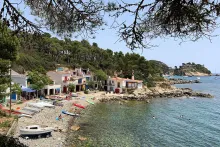 Wide shot of a quaint but beautiful seaside village, with a thin strip of rocky beach, lined by small boats, separating homes and trees from the Mediterranean Sea.