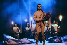 A woman stands on stage, holding a hand drum and a beater stick. Five actors behind her hold up torches. They all wearing matching white outfits with mock bandoliers. The stage is foggy and lit blue.