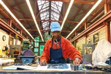 A woman reaches across a table while standing in a studio wearing a blue hat, an orange collared shirt, and a blue smock. Behind the woman are studio materials.