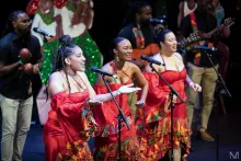 Three women singers in matching red dresses perform on stage, with male musicians and Christmas decorations on stage behind them.