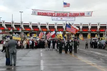 People dressed in military uniform and formal wear cluster at the start of a parade route on a wide road. In the background, an American flag flying above a banner that reads 'Bienvenidos Washington's Birthday Celebration,' and a solid line of inspection booths at an international border.