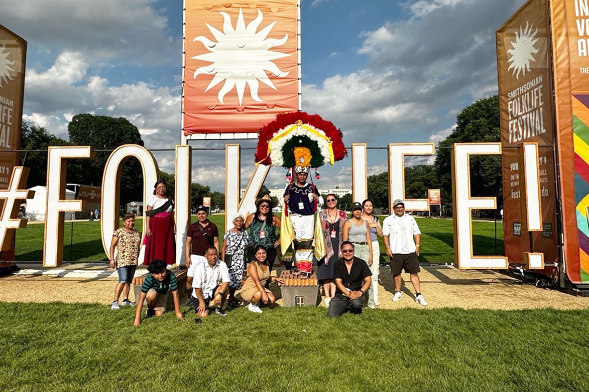 Fourteen people pose outdoors in front of a large marquee reading #FOLKLIFE and underneath an orange banner of the Smithsonian sunburst logo. A person near the center wears a large, semicircular feather headdress in red, white, green, and yellow.